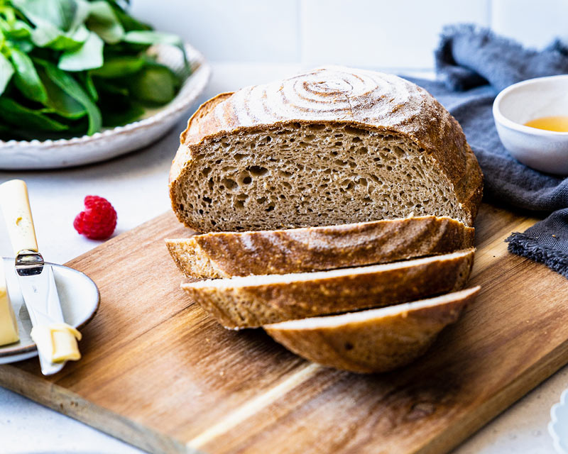 Pain Piaf sans gluten coupé en tranches sur une planche en bois, mie alvéolée, salade verte et couteau à beurre